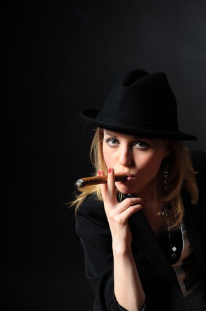Beautiful girl in a hat with a cigar on a black background looks into the cameraの写真素材