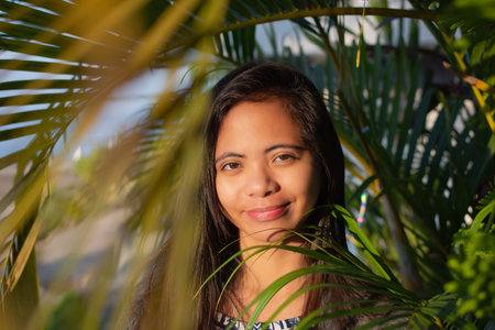 portrait of a beautiful young Filipina woman against a background of palm leaves in the rays of the sunの写真素材