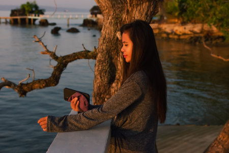 a young Asian woman stands alone on the seashore with a phone in her hands and looks into the distance.の写真素材