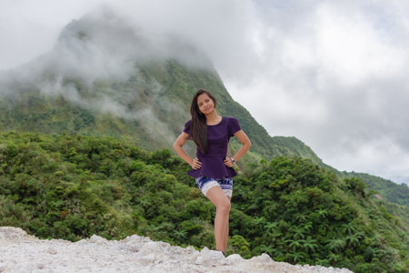 Portrait of a beautiful young Filipina woman against the background of a volcano covered with smog and cloudsの写真素材