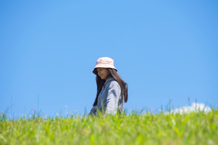Young Asian woman walking on a green hill, smiling at the camera, against a blue sky.の写真素材
