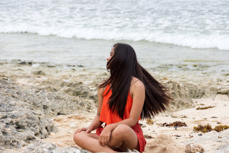 Young woman with flowing long brunette hair in swimsuit sitting on rocky beach by the seaの写真素材