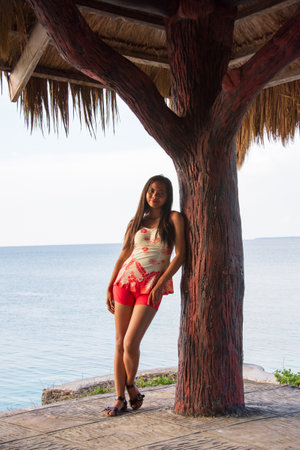 young asian woman in one piece swimsuit posing near tree in hut by seaの写真素材