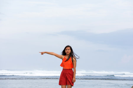 Young Asian woman in an orange bikini top and red skirt points her hand towards the sea while standing on the shoreの写真素材