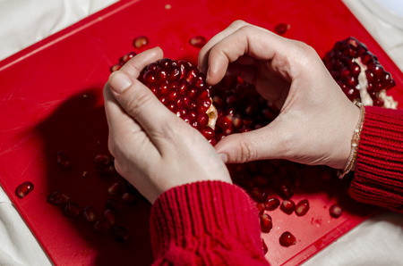 Human hands making a heart of fresh pomegranate pices on the red cutting board. The background is red. Ripe organic food close up. Stay healthy concept. Broken symbol of love.の写真素材