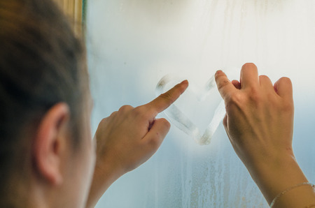Woman hands drawing a heart on the sweaty foggy window. Water droplets condensation background of dew on glass, symbol of love close up. Be happy and love even in bad raining weather outside concept.の写真素材
