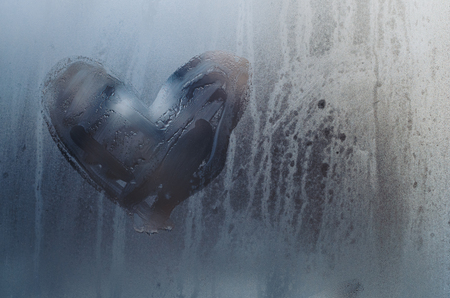A heart is drawn on the sweaty foggy window. Water droplets condensation background of dew on glass, symbol of love close up view. Be happy and love even in bad raining weather outside concept.の写真素材