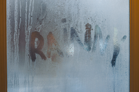 A word Rainy is written on the sweaty foggy window. Water droplets condensation background of dew on glass. Be happy even in bad rainy weather outside concept.の写真素材