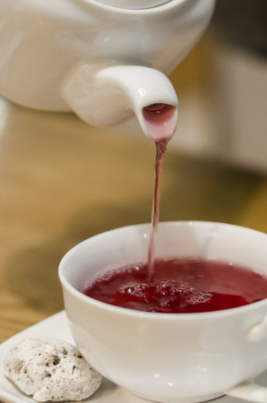 Pouring fruit red tea from the teapot into white cup concept. Eating breakfast on the wooden table background. Dinking a hot drink in the coffee shop.の写真素材