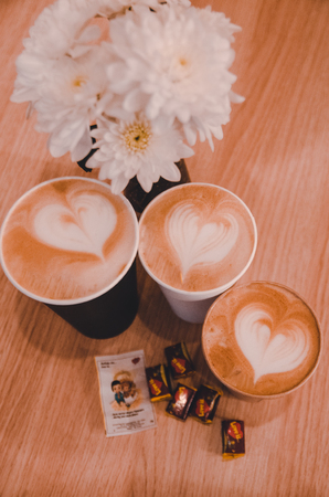 A gift for your couple on Valentines day concept. Latte art with a symbol of love. Coffee in takeaway cups, "love is..." gum and white flowers in the transparent vase with water on the wooden table.の写真素材