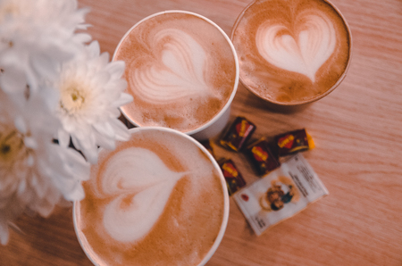 A gift for your couple on Valentines day concept. Latte art with a symbol of love. Coffee in takeaway cups, "love is..." gum and white flowers in the transparent vase with water on the wooden table.の写真素材