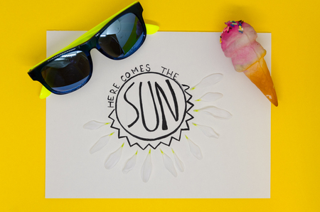White page of notebook where is written with nice handwriting: Here comes the sun, isolated on yellow background. A greeting to spring concept. White flower leaves, sunglasses and icecream as a decorの写真素材