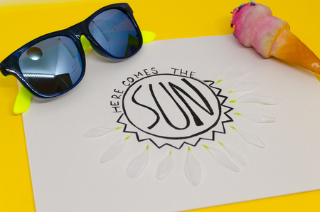 White page of notebook where is written with nice handwriting: Here comes the sun, isolated on yellow background. A greeting to spring concept. White flower leaves, sunglasses and icecream as a decorの写真素材