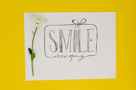 White page of a notebook where is written with nice handwriting: Smile its spring, isolated on yellow background. Hello spring greeting concept. A gift as a frame and a flower as a decorの写真素材