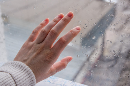 a hand touching the glass window with rain drops. Woman fingers on the window background. Rainy and cold weather outside. View on the street from the room.の写真素材