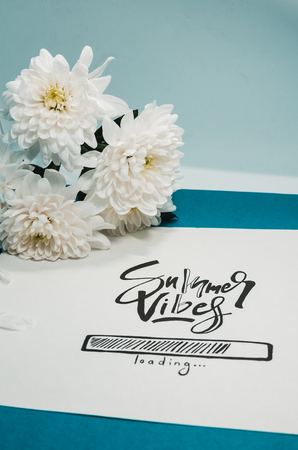 Hello summer concept.  A white paper where is wtitten with nice handwriting - Summer vibes loading. Summer quote and a white flower on the blue backgroundの写真素材