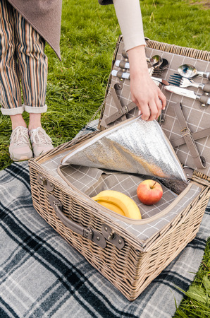 woman opening picnic basket with food and silver cutlery on the grey blanket on the grass. bananas and apples in wicker hamper.の写真素材
