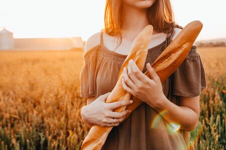 woman holds freshmade french baguette in wheat field during sunrise in countrysideの写真素材