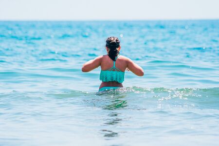 woman in blue swimsuit walks into black sea from the back viewの写真素材