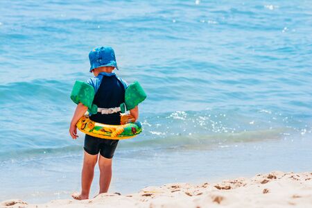a child in the swimming circle and arm rings is walking on the sand beach to swim in the sea.の写真素材