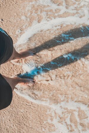 Close-up of a man's bare feet walking on the beach , with the edge of the wave foaming softly under...