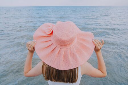 a girl in a white dress holds in her hands a large pink hat and looks towards the sea. Dreams Come True Concept .の写真素材