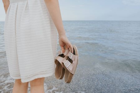 woman in a white dress holds beach slippers in her hands and walks along the shore of the blue sea