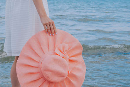woman in a white dress holds pink hat along the blue seaの写真素材