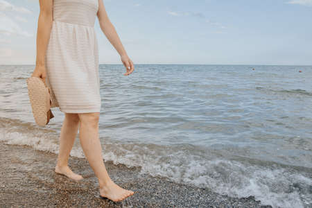woman in a white dress holds beach slippers in her hands and walks along the shore of the blue seaの写真素材