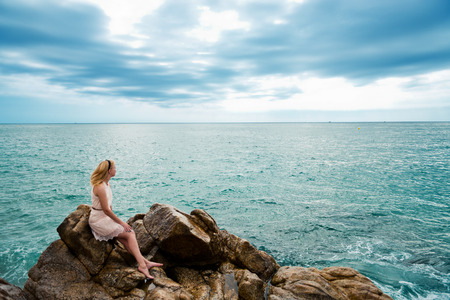 Beautiful young woman sitting on the stone on the seashoreの写真素材