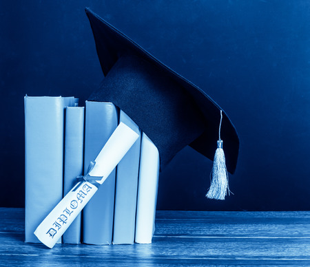 A mortarboard and graduation scroll, tied with red ribbon, on a stack of booksの写真素材