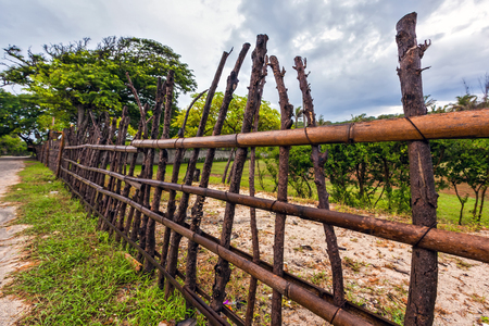 A fence made of bamboo wood and brown twisted curves on the background of a littered yard with green trees in cloudy weather. Branch fence closeup, Gili Trawangan, Indonesia.の写真素材