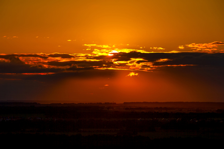 Dark orange sunset, the sun disc hides behind the clouds in the middle of the frame, casts shadows on the valley with silhouettes of fields and forests.の写真素材