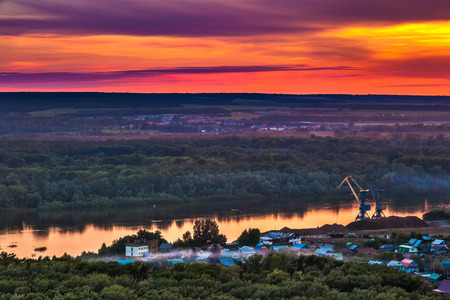 Bright red yellow orange setting sun on a blue sky with dramatic dark clouds over the city on the banks of the Belaya River in a green dense forest.の写真素材