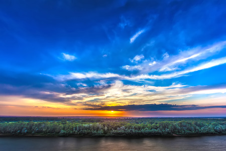 Incredibly beautiful colorful sunset on the river bank, bright orange sun hidden behind white clouds on a saturated blue sky above the green forest.の写真素材