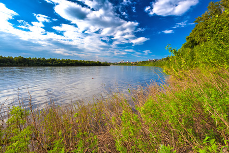 Belaya river bank, overgrown with lush shrubs and green trees under colorful white clouds against...