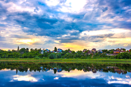 The quiet blue water with the reflection of the sky with massive low clouds, yellow rays of the sun above the green overgrown shore, private country houses.の写真素材