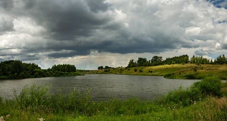 Cloudy windy summer landscape with river and green hills.