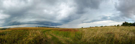Summer cloudy panoramic landscape with rural road passing through meadows and fields. Ominous clouds in overcast sky over the ripe wheat agricultural fields. Heavy rain and thunderstorm coming.の写真素材