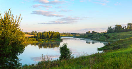 Summer serene landscape with calm river and green hills at sunrise.