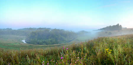 Beautiful summer calm hazy landscape with foggy hollow and green hills before the sunrise.Calm forest river flowing between the woods and meadows with blooming wildflowers during misty morning.の写真素材