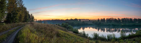 Beautiful panoramic landscape with rural dirt road and calm lake at sunrise.Colorful fiery sky over the green hills and woods.の写真素材