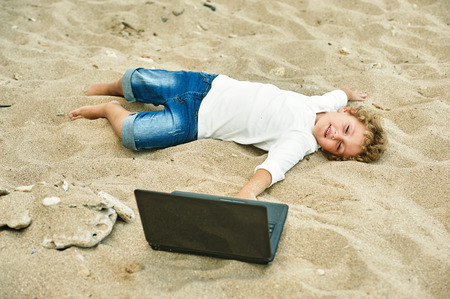 a boy plays on the beach, lying next to the laptopの写真素材