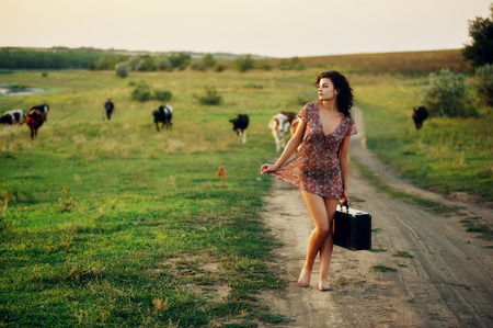 Beautiful young model girl in a summer dress in a field on a dirt road . The concept of rustic retro style clothingの写真素材
