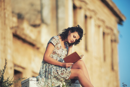 A young woman in a light summer dress reading book outdoorsの写真素材