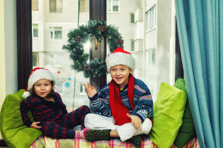 Funny kids waiting for Christmas . Brother and sister on the window dressed in Santa hatsの写真素材