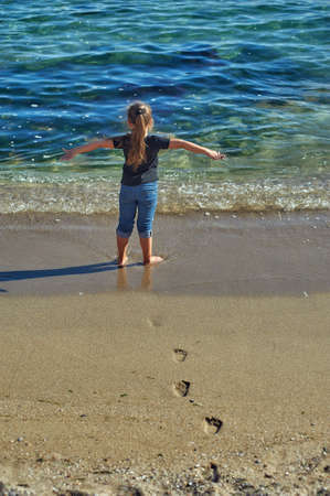 A little girl dressed in blue jeans and a t-shirt standing with her back to the sea.の写真素材