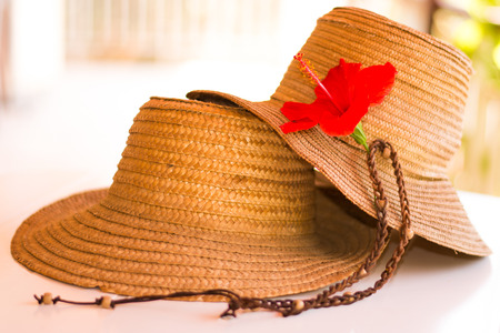 Two straw hats on a white table, with an hibiscus flower on one of them.