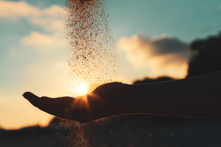 Silhouette of a young girl's hand pouring sand from a beach against a blue sky at sunsetの写真素材