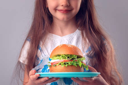 Little girl eating big burger. Kid looking at healthy big sandwich, studio isolated on white background.の写真素材